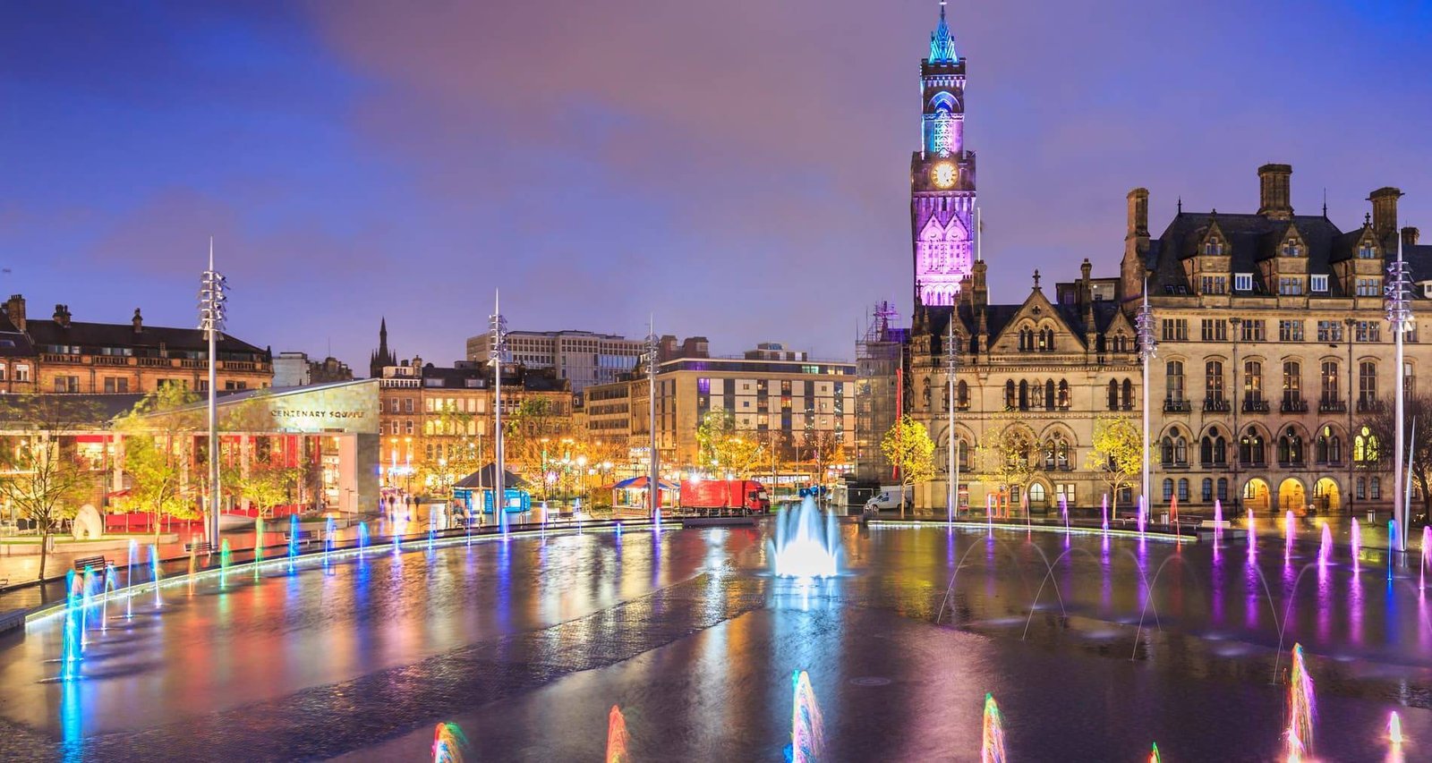 Night view of a the bradford city centenary square with lit fountains, buildings, and a clock tower with colorful lights reflecting on the water.